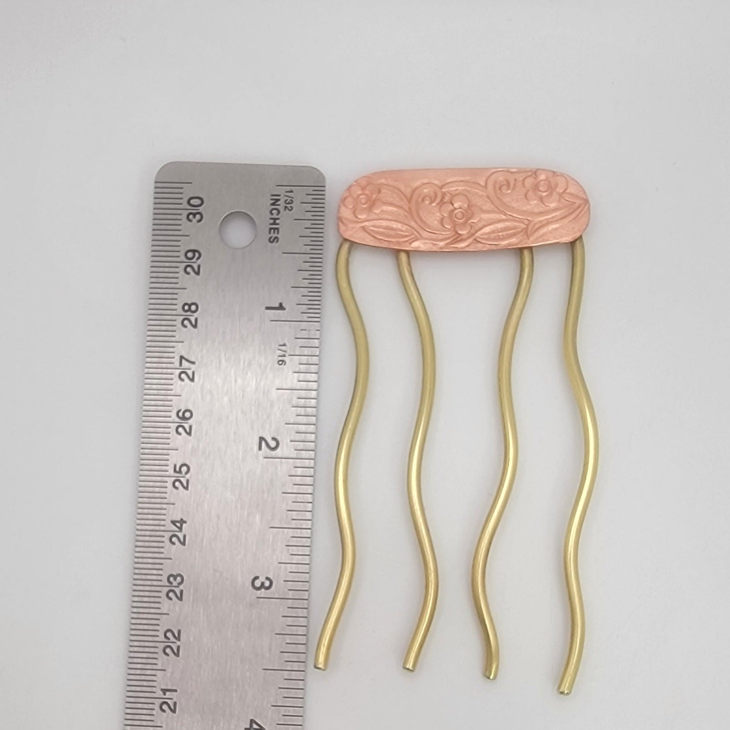Brass hairpin with floral design on a copper plate next to a ruler for scale on a gray background