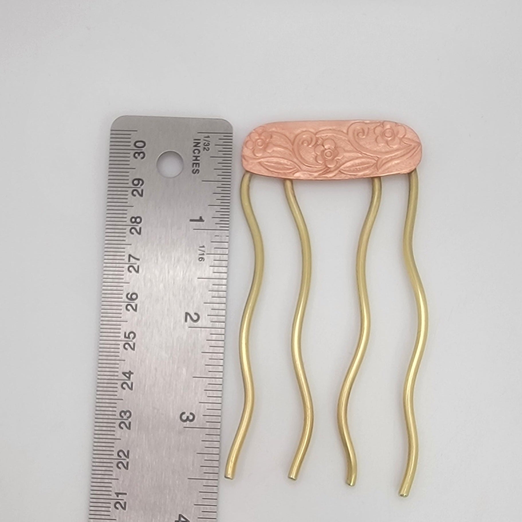 Brass hairpin with floral design on a copper plate next to a ruler for scale on a gray background