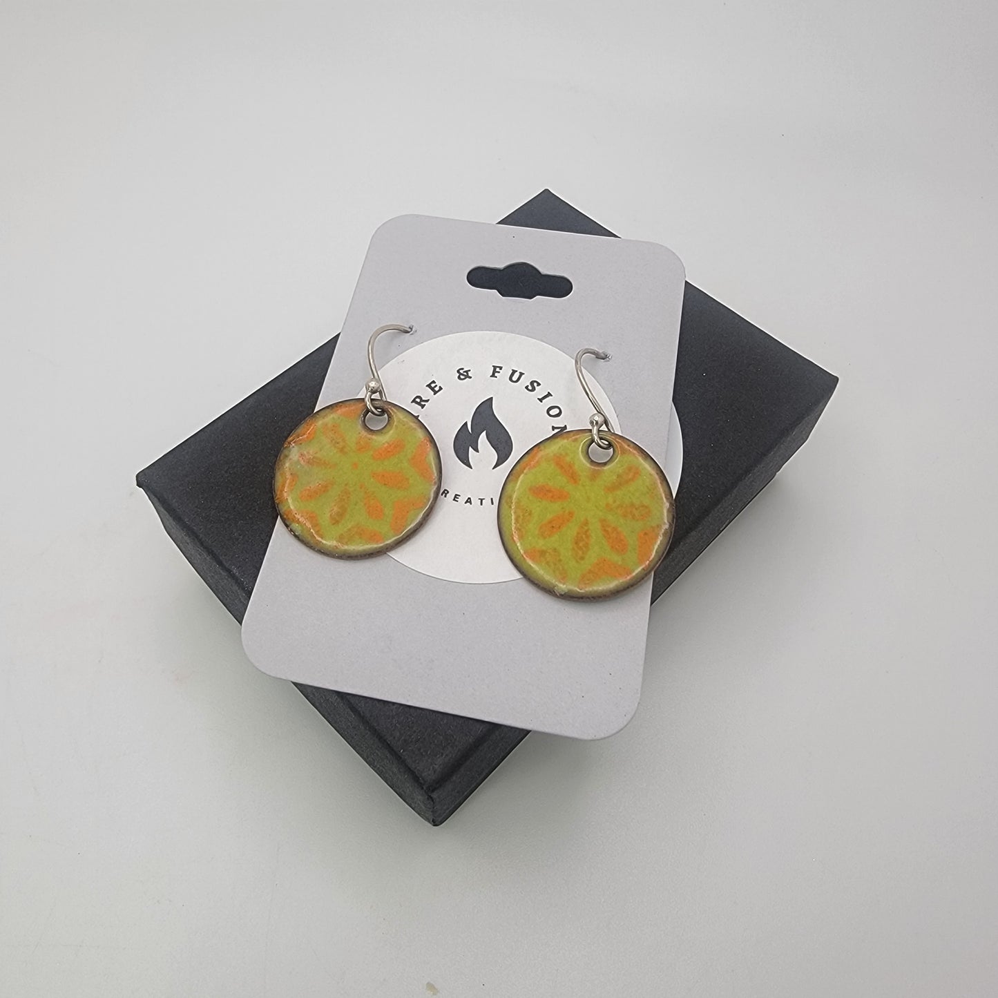 Pair of round earrings with orange and green design on a branded card against a white background