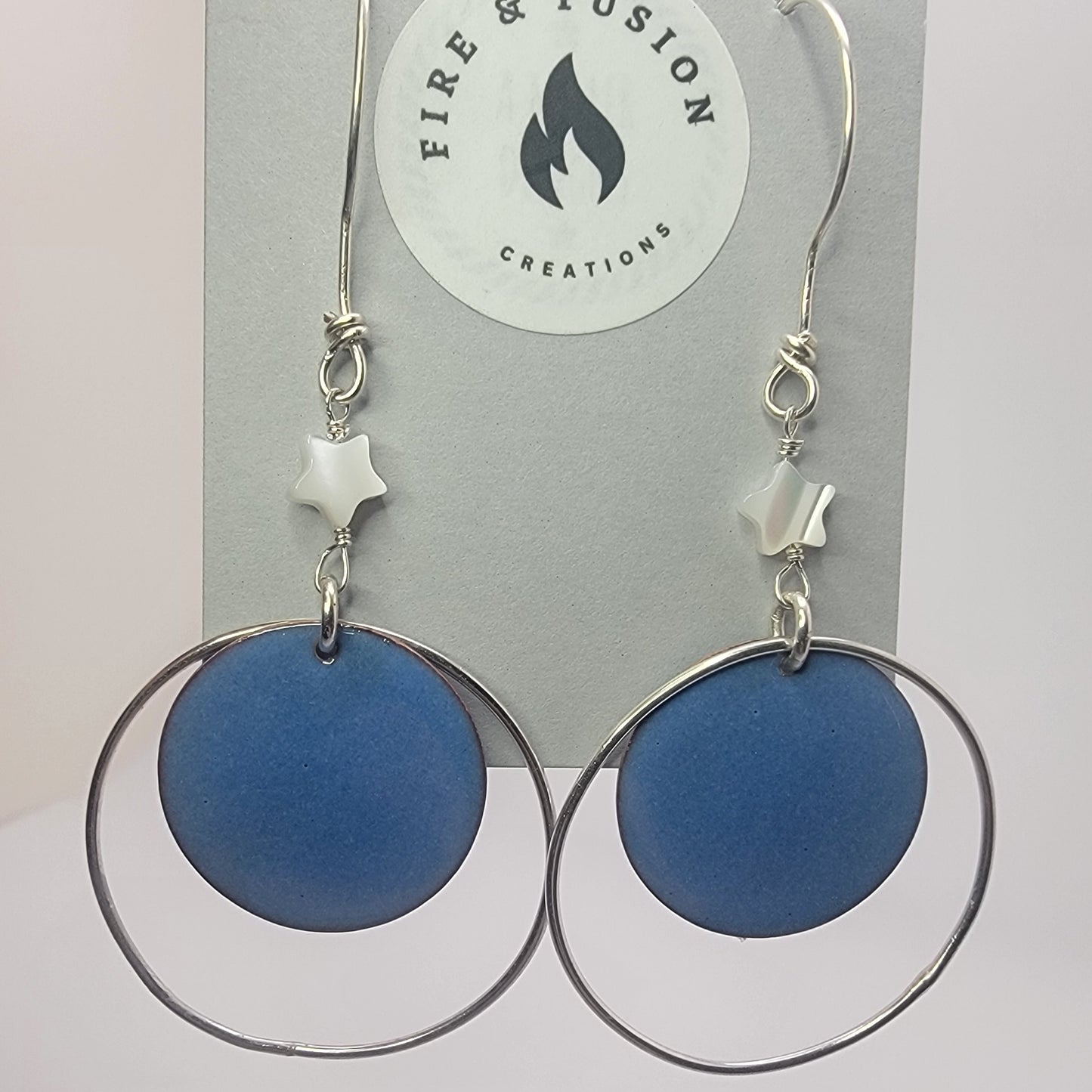 Blue circular earrings with silver hoops on jewelry card titled ' Fire and Fusion Creations' against white background