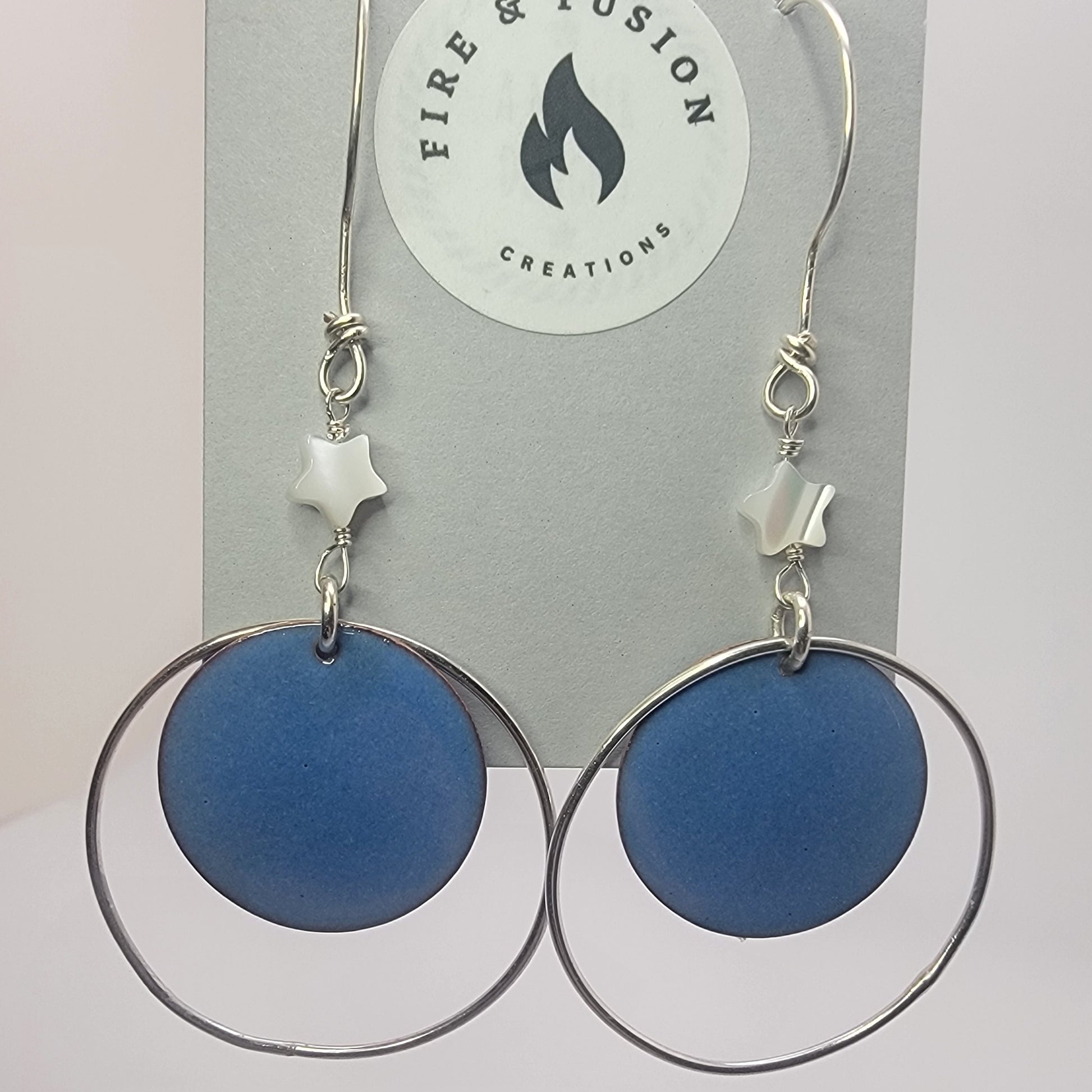 Blue circular earrings with silver hoops on jewelry card titled ' Fire and Fusion Creations' against white background