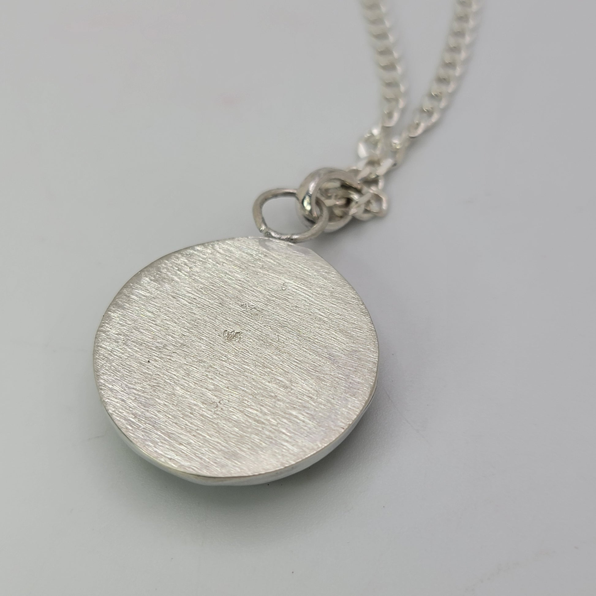 A silver round disc pendant necklace with a brushed texture, hanging from a silver chain on a light gray background.