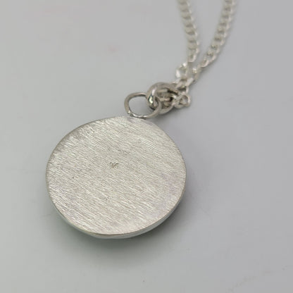 A silver round disc pendant necklace with a brushed texture, hanging from a silver chain on a light gray background.