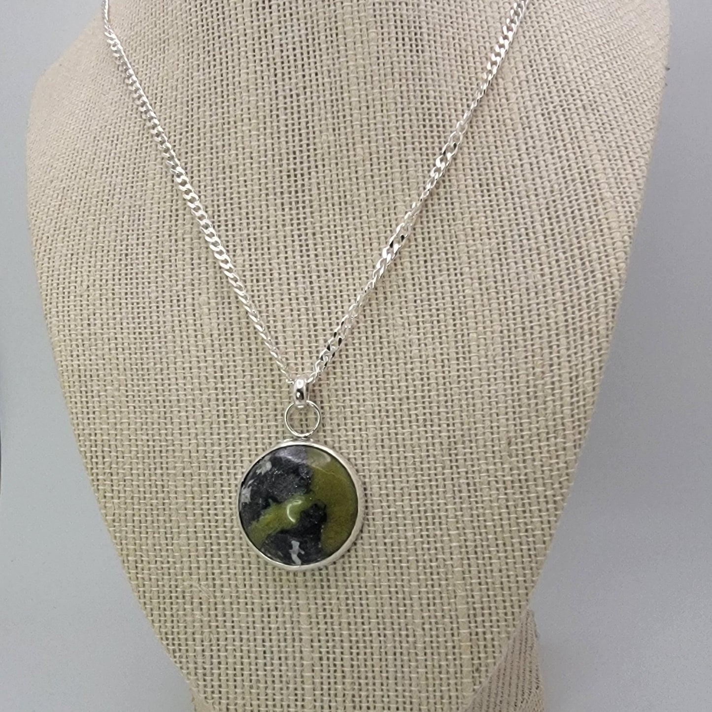 A silver chain necklace with a round pendant featuring a green and black marbled pattern, displayed on a beige fabric jewelry bust.