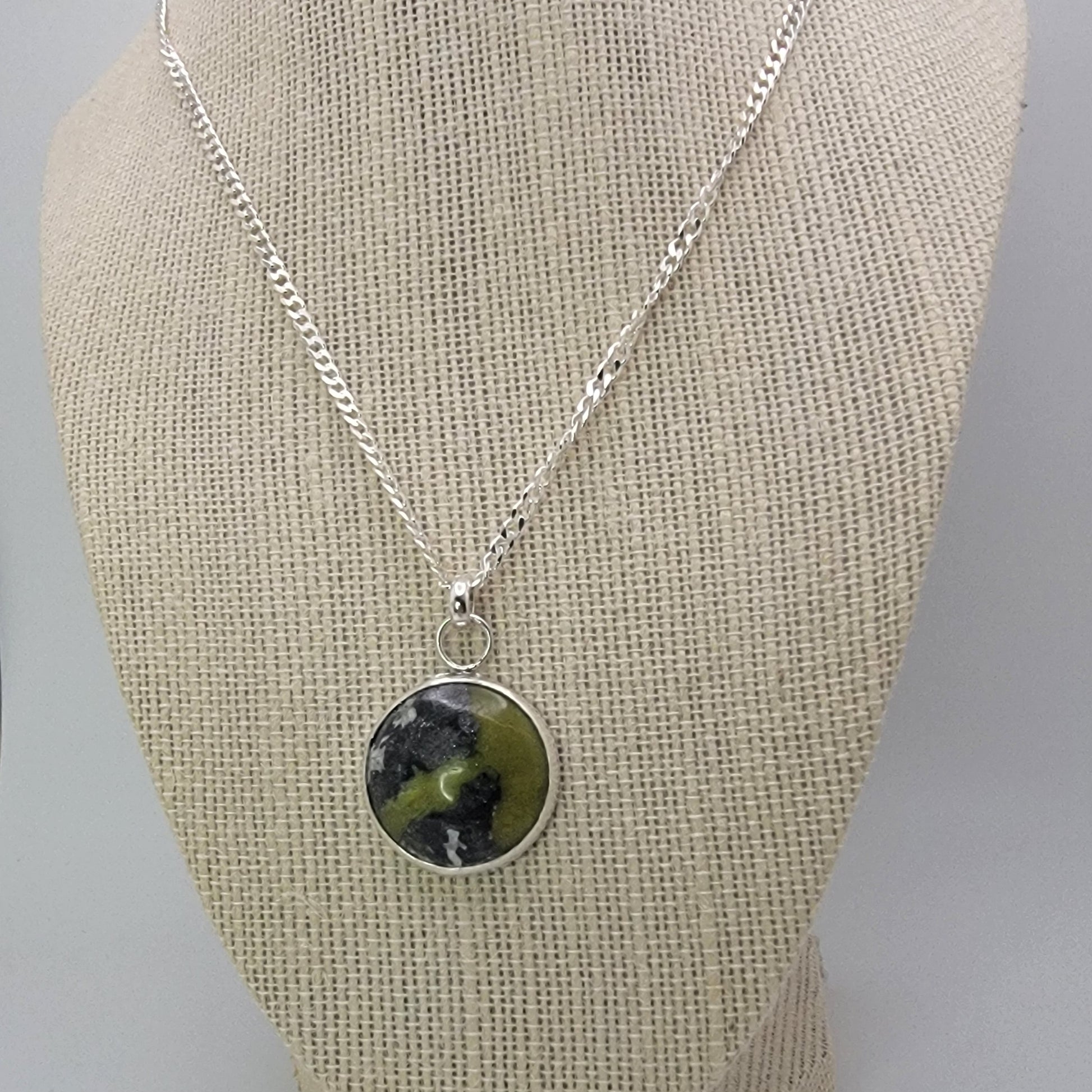 A silver chain necklace with a round pendant featuring a green and black marbled pattern, displayed on a beige fabric jewelry bust.