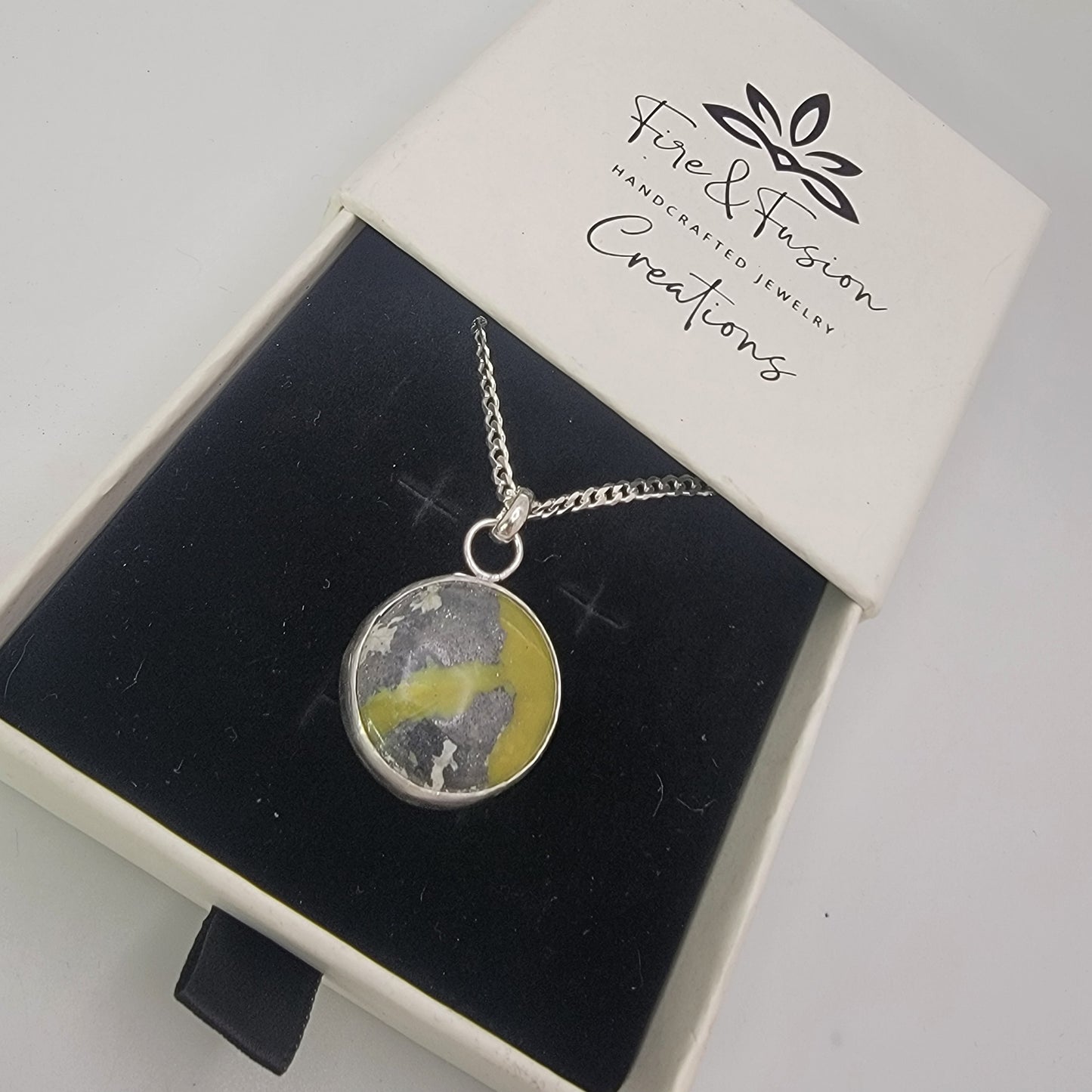 A silver chain necklace with a round pendant containing a yellow and gray marbled pattern, presented in a white jewelry box with a black velvet insert. The brand 'Fire & Fusion Creations' is printed on the box lid.