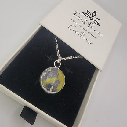 A silver chain necklace with a round pendant containing a yellow and gray marbled pattern, presented in a white jewelry box with a black velvet insert. The brand 'Fire & Fusion Creations' is printed on the box lid.