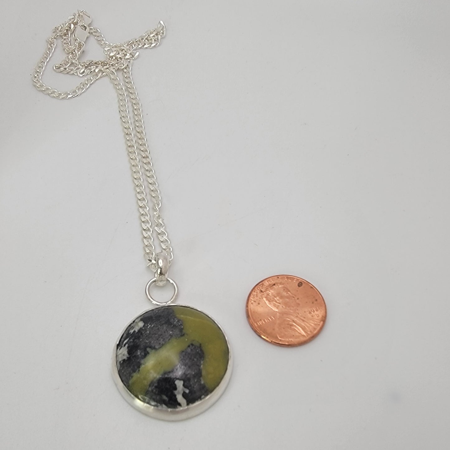 A silver chain necklace with a round pendant featuring a black, white, and olive-green marbled stone, displayed next to a copper penny for scale on a light gray background.