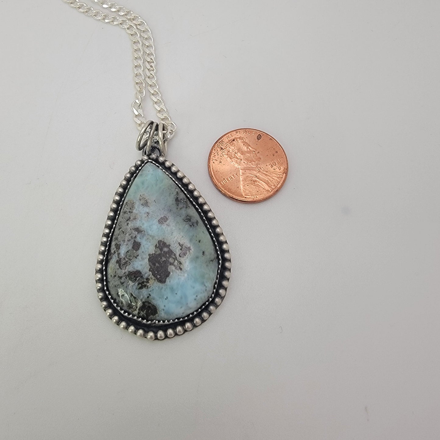 Blue gemstone and bezel set silver pendant on silver curb chain next to penny for scale on white background