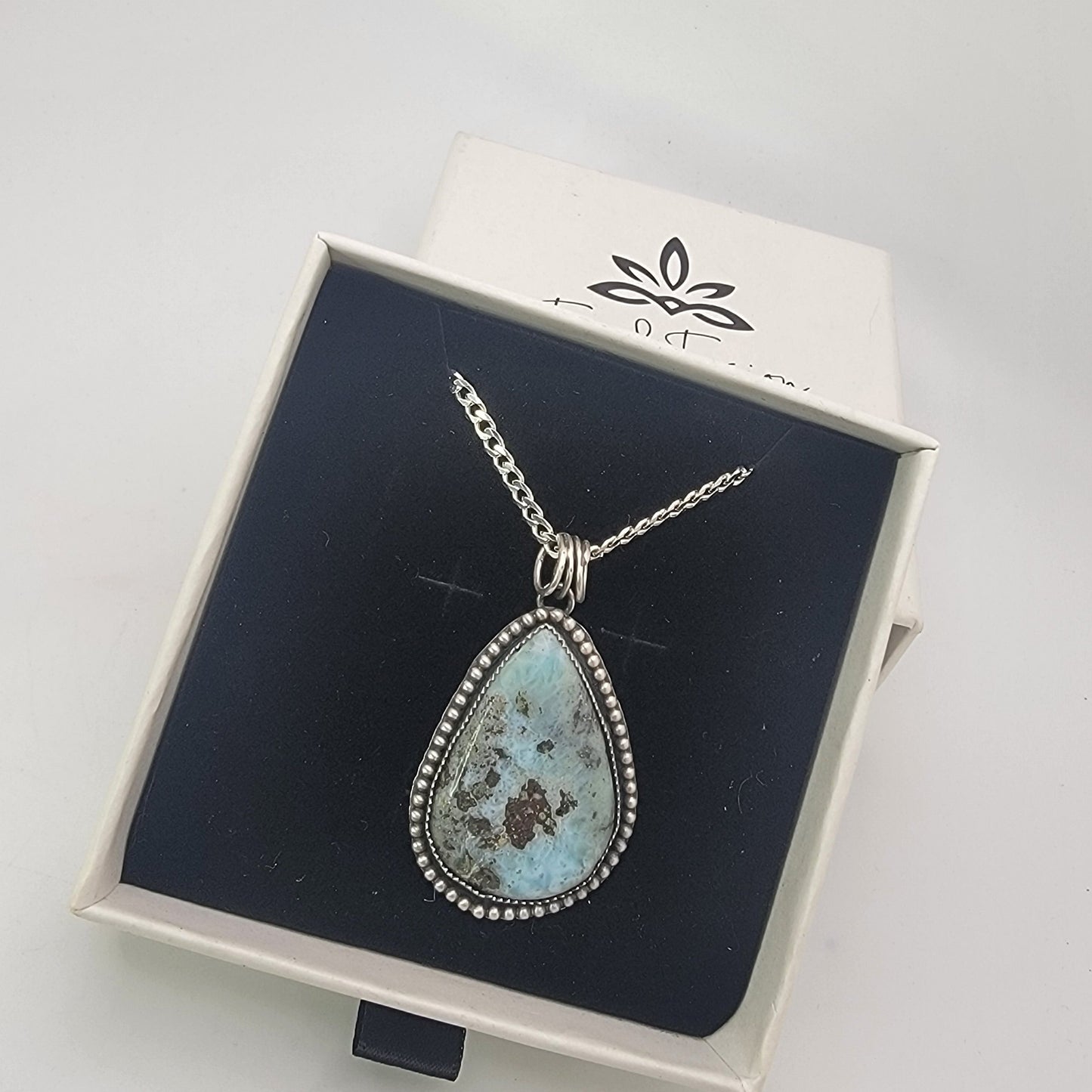 Blue teardop shaped gemstone bezel set pendant shown in branded white gift box on black interior