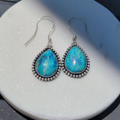 Pair of blue gemstone earrings bezel set in silver with oxidized beaded accent on silver hooks on a white background