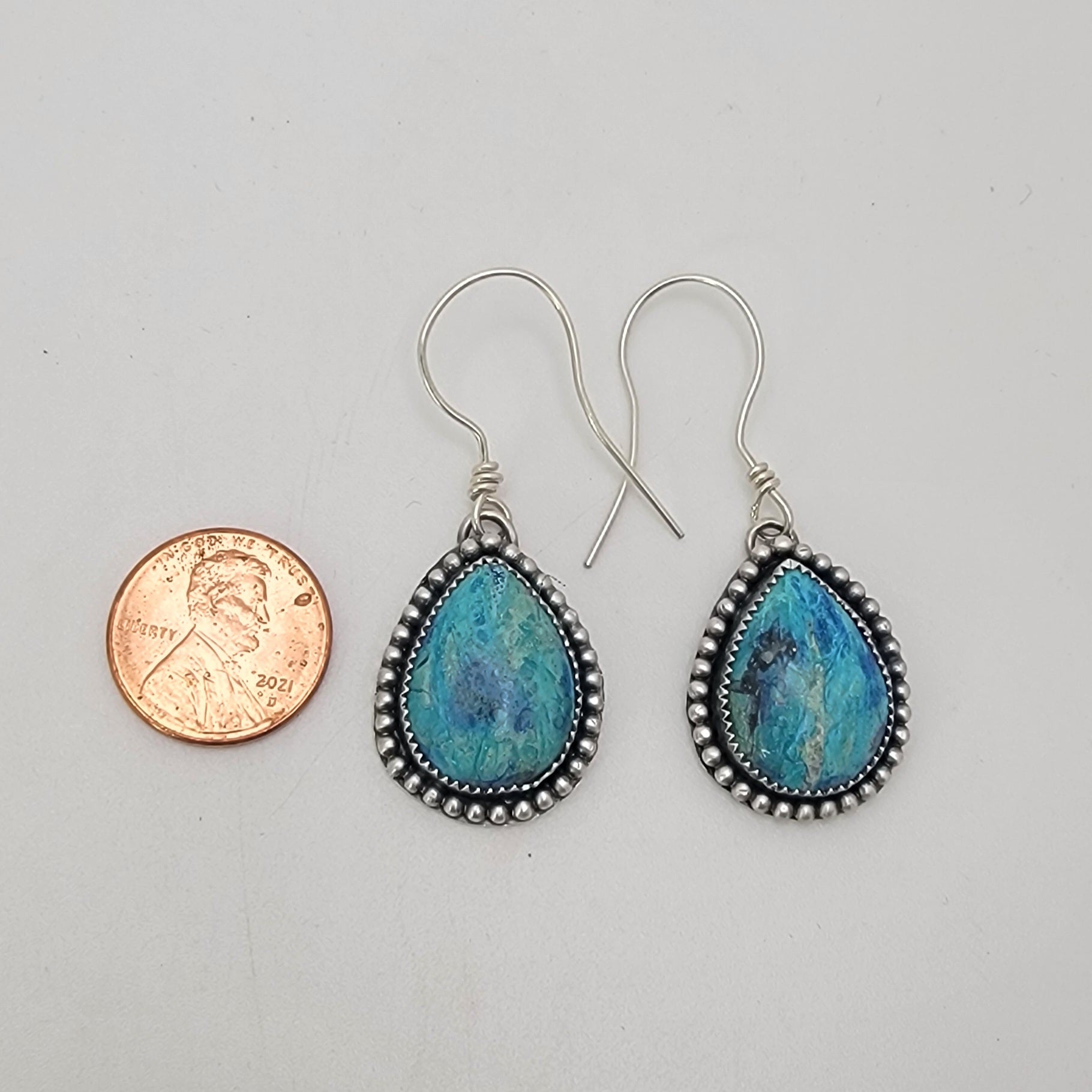Pair of blue gemstone earrings bezel set in silver with oxidized beaded accent on silver hooks on a white background next to a penny for scale
