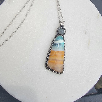 A silver-toned pendant necklace featuring a teardrop-shaped stone with horizontal bands of turquoise, cream, and orange, displayed on a textured white circular surface.