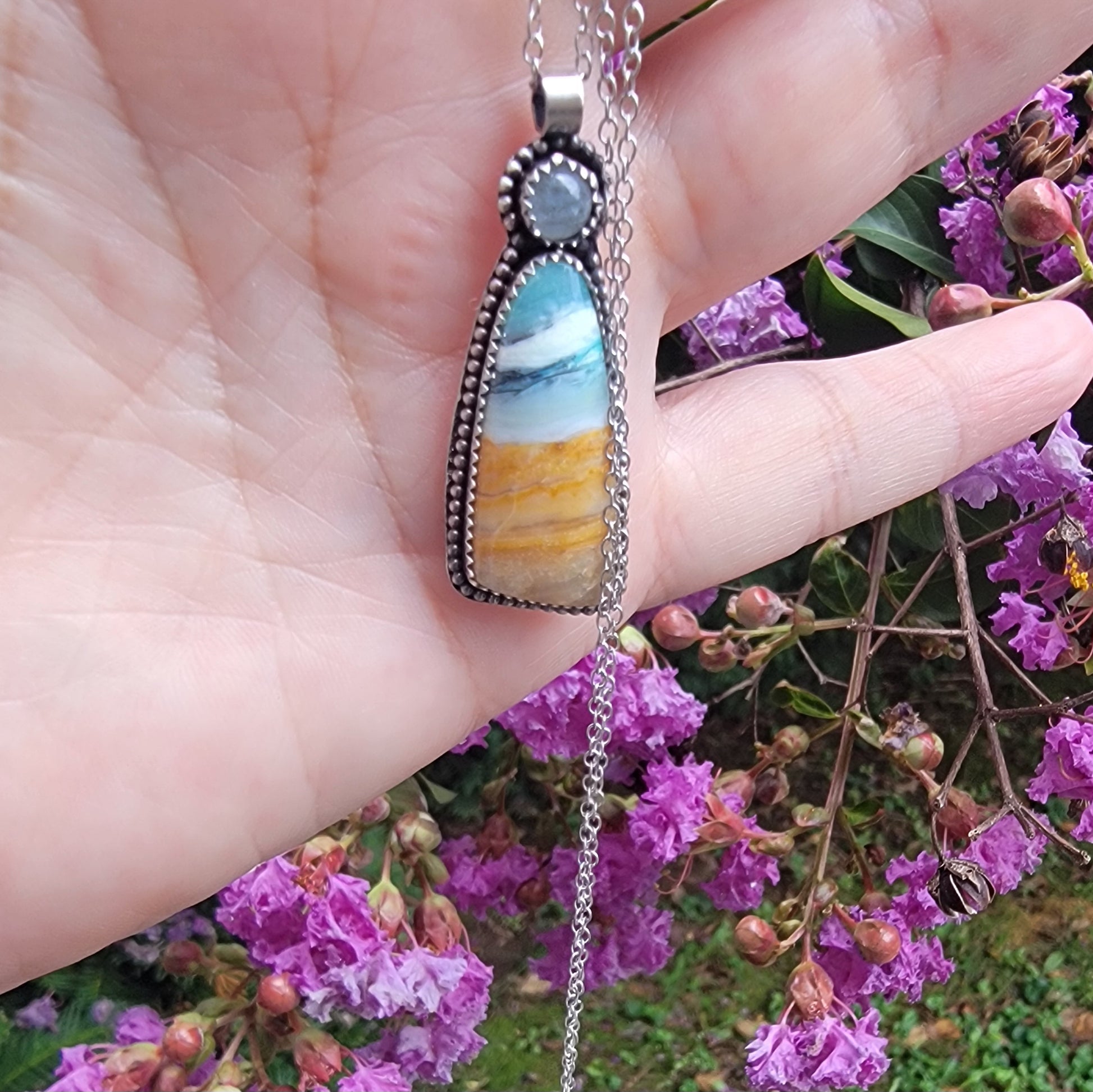 A silver-toned pendant necklace with a layered stone featuring blue, white, and golden-yellow bands, held in a person's hand against a background of purple flowers.