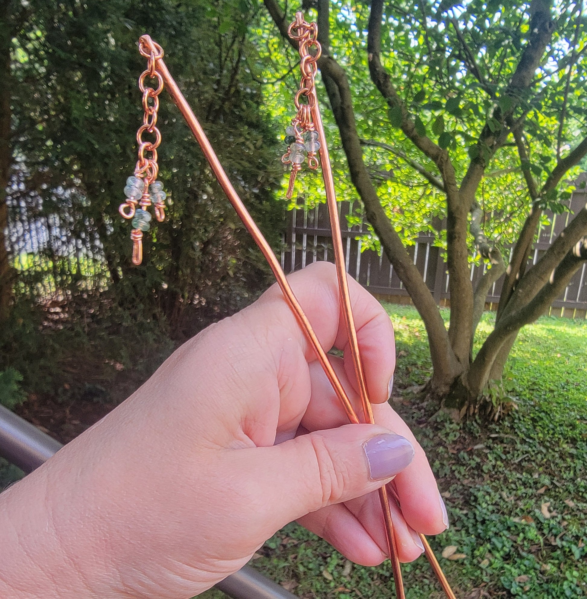Hand holding a pair of copper beaded hair sticks against an outdoor natural background