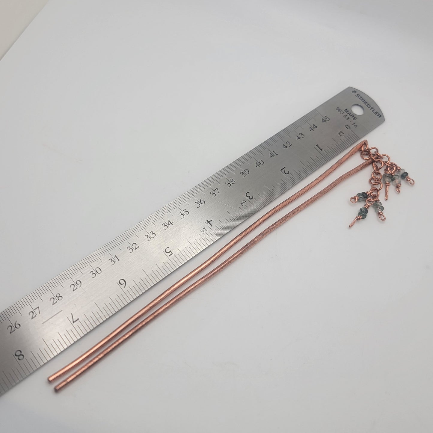 A pair of copper beaded hair sticks next to a metal ruler on a white background