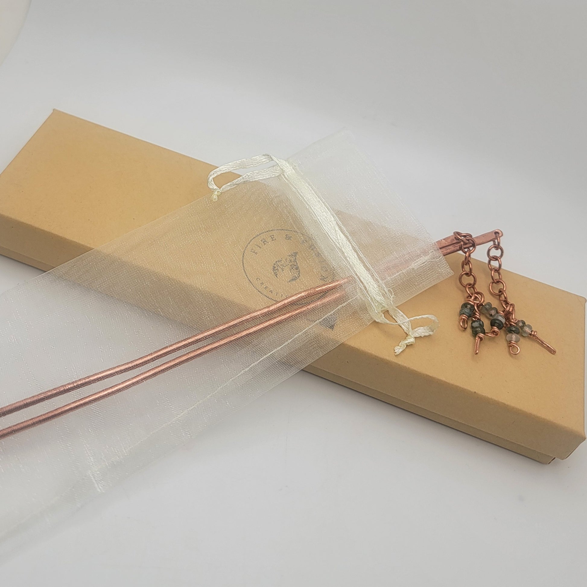 A pair of copper beaded hair sticks in an organza gift bag on a long cardboard gift box on a white background