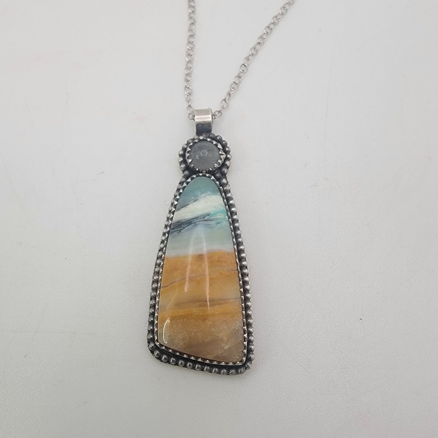 A silver pendant necklace featuring a teardrop-shaped stone with a marbled pattern of blue, white, and golden-brown, suspended from a silver chain against a plain white background.