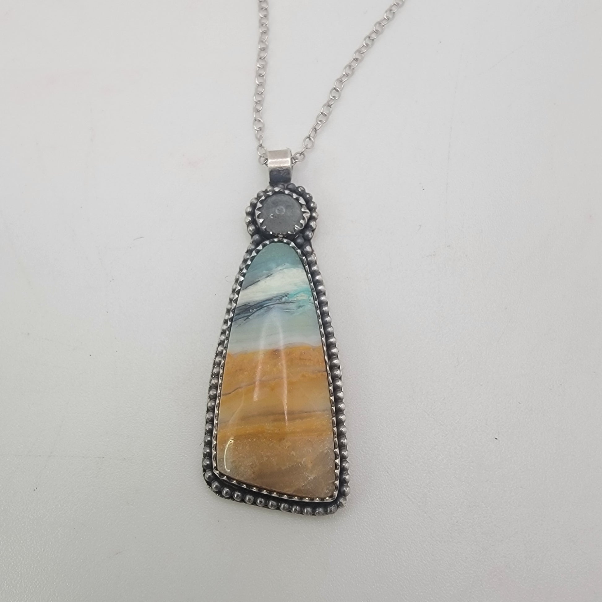 A silver pendant necklace featuring a teardrop-shaped stone with a marbled pattern of blue, white, and golden-brown, suspended from a silver chain against a plain white background.