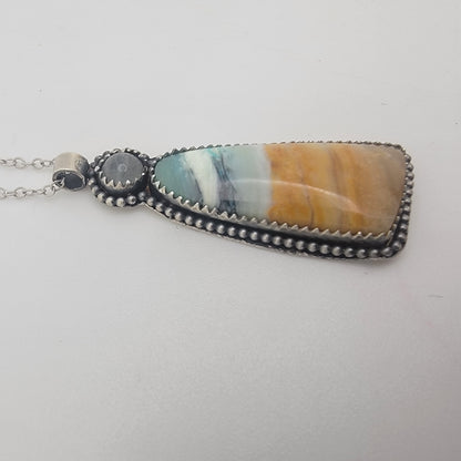 A silver-toned beaded pendant necklace featuring a large, teardrop-shaped cabochon stone with horizontal bands of light blue, white, and warm orange-brown, displayed on a neutral light gray background.