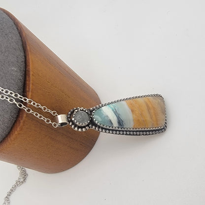 A silver-toned pendant necklace with a teardrop-shaped stone featuring blue, white, and golden-brown marbled patterns, displayed on a wooden jewelry stand against a white background.