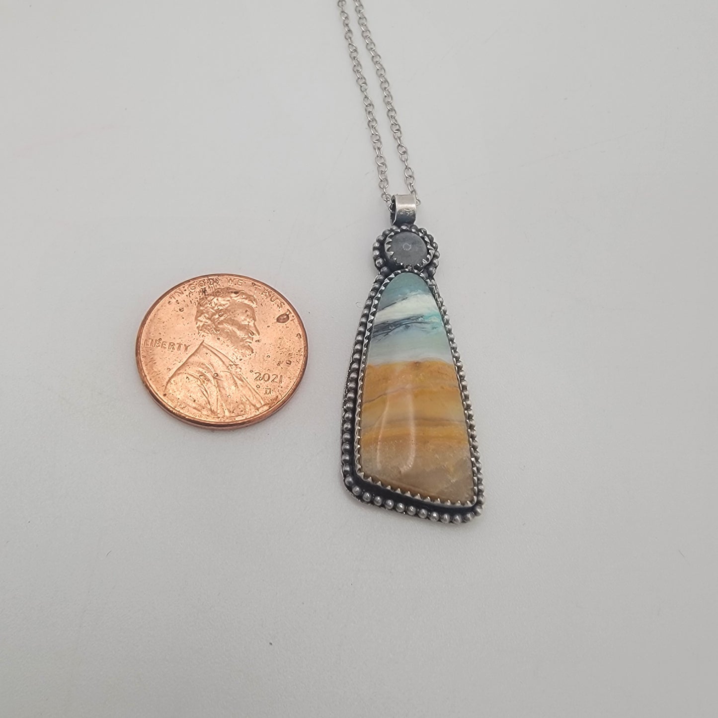 A silver beaded pendant necklace with a triangular stone featuring a blue, white, and orange marbled pattern, shown next to a US penny for scale on a light gray background.