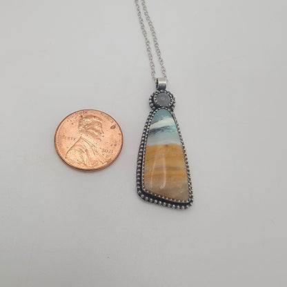 A silver beaded pendant necklace with a triangular stone featuring a blue, white, and orange marbled pattern, shown next to a US penny for scale on a light gray background.
