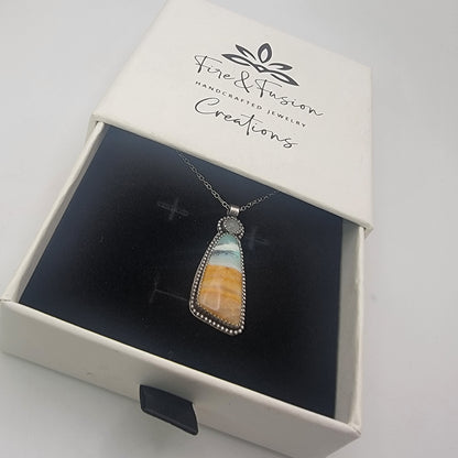 A silver-toned pendant necklace with a triangular stone showing blue, white, and orange banding, displayed inside an open white jewelry box branded 'Fire & Fusion Handcrafted Jewelry Creations'.