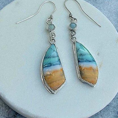A pair of silver dangle earrings with teardrop-shaped turquoise and orange stone pendants, displayed on a round white surface.