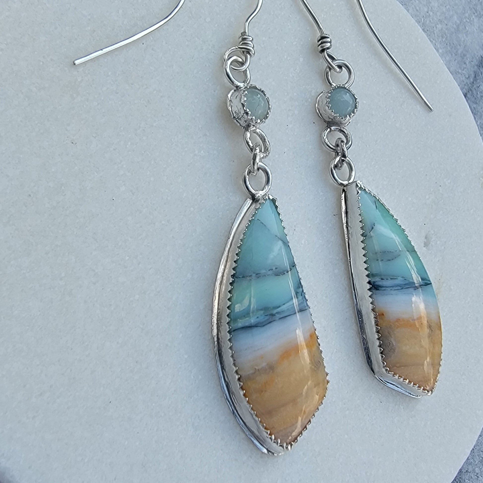 A pair of silver dangle earrings with teardrop-shaped turquoise and amber marbled stone pendants, displayed on a light gray textured surface.