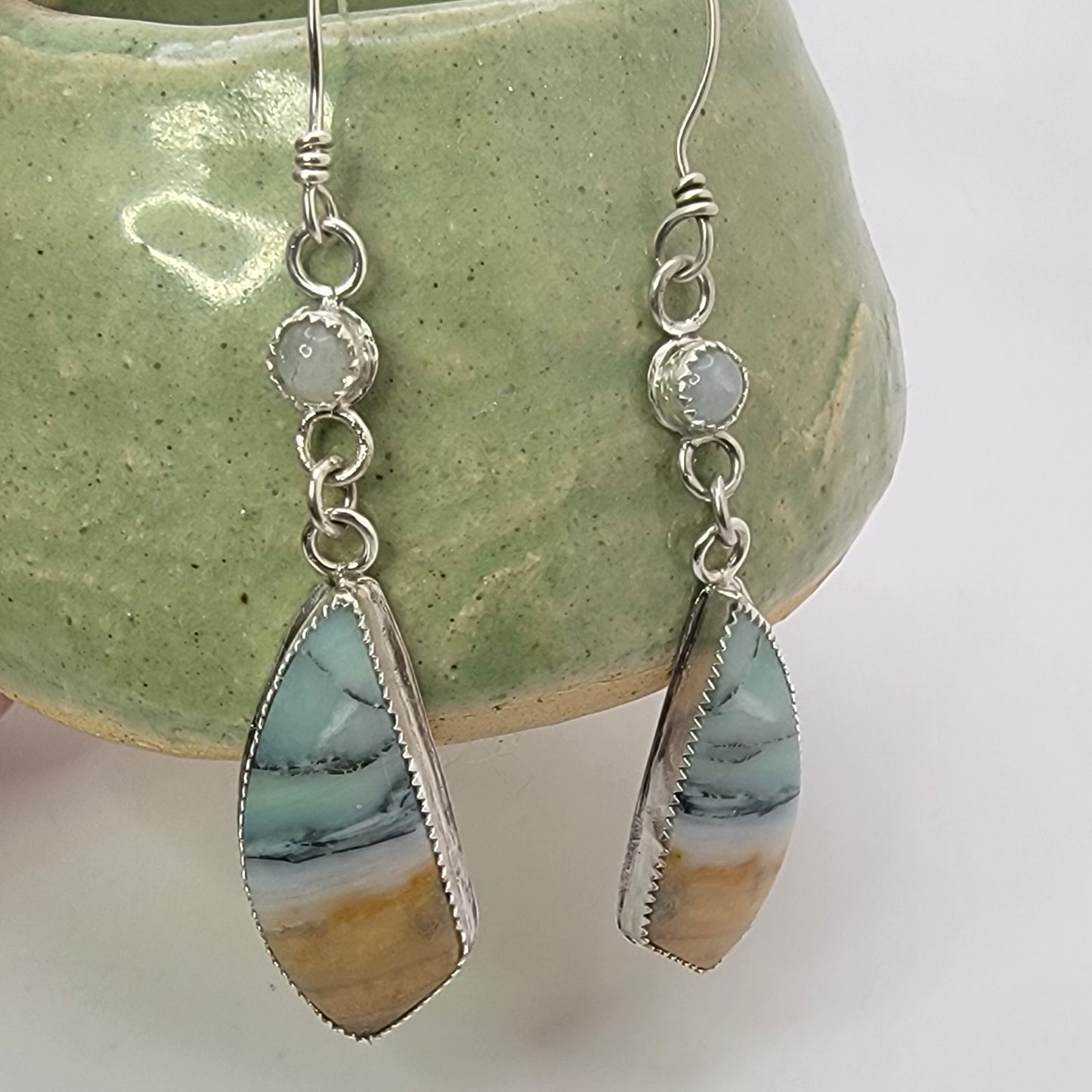 A pair of silver dangle earrings with teardrop-shaped marbled stone pendants in shades of blue, white, and tan, displayed against a light green ceramic bowl on a white background.