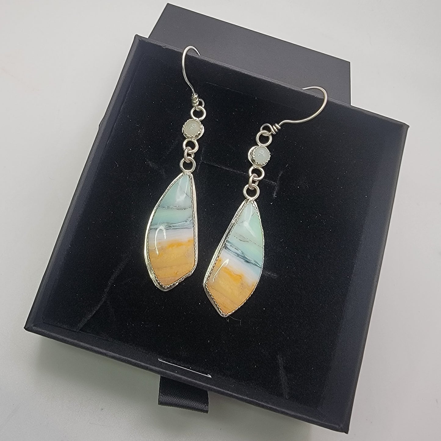 A pair of silver-toned dangle earrings with teardrop-shaped marbled stone pendants in white, light blue, and orange-yellow, displayed on a black presentation box.