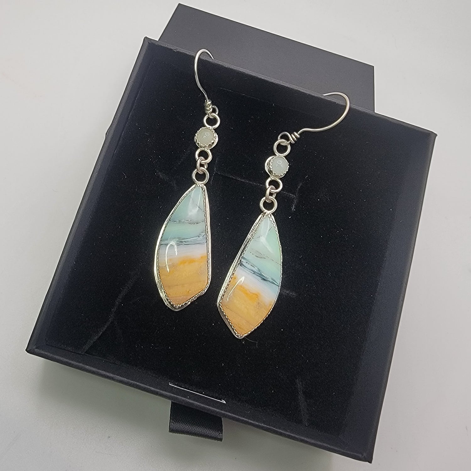 A pair of silver-toned dangle earrings with teardrop-shaped marbled stone pendants in white, light blue, and orange-yellow, displayed on a black presentation box.