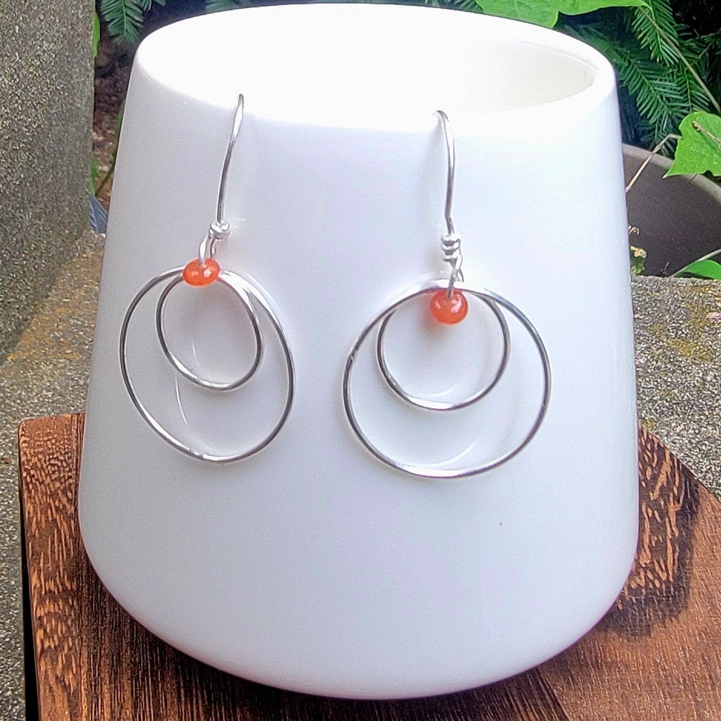 A pair of silver double-ring hoop dangle earrings with small orange beads, displayed on a white ceramic cup.