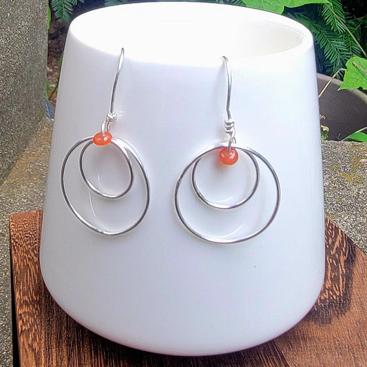 A pair of silver double-ring hoop dangle earrings with small orange beads, displayed on a white ceramic cup.