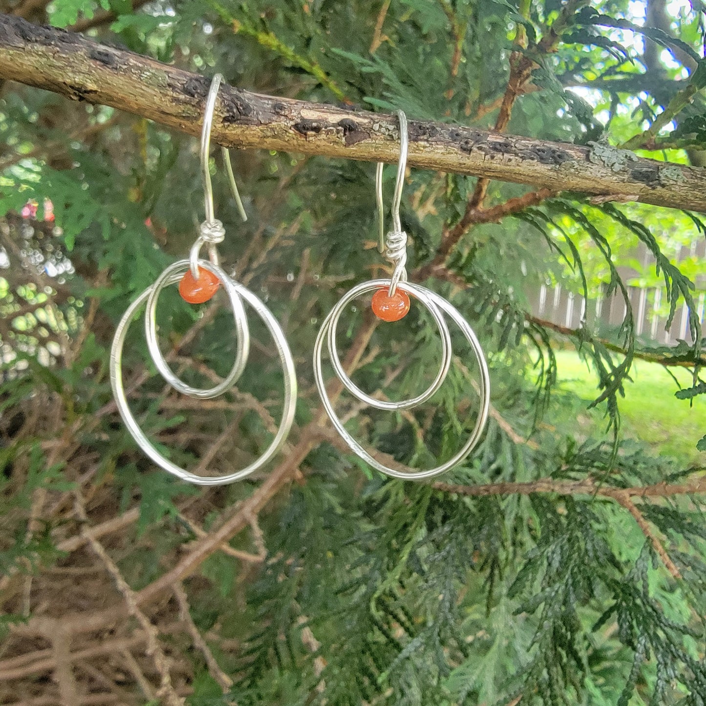 A pair of silver hoop earrings with small orange bead accents, hanging from a tree branch with green foliage in the background.