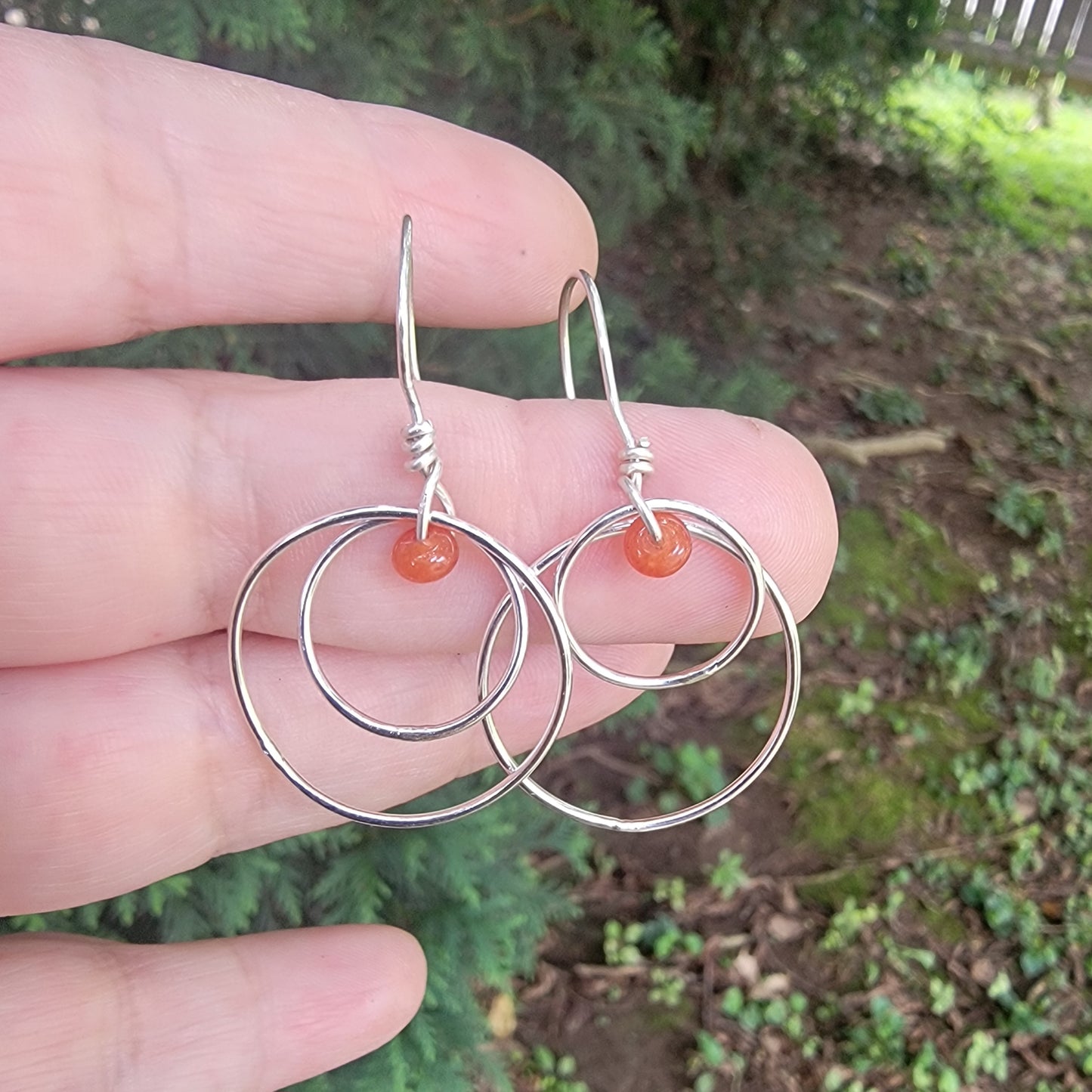 A pair of silver hoop earrings with small orange bead accents, held in a person's hand against a blurred outdoor background of greenery and mulch.