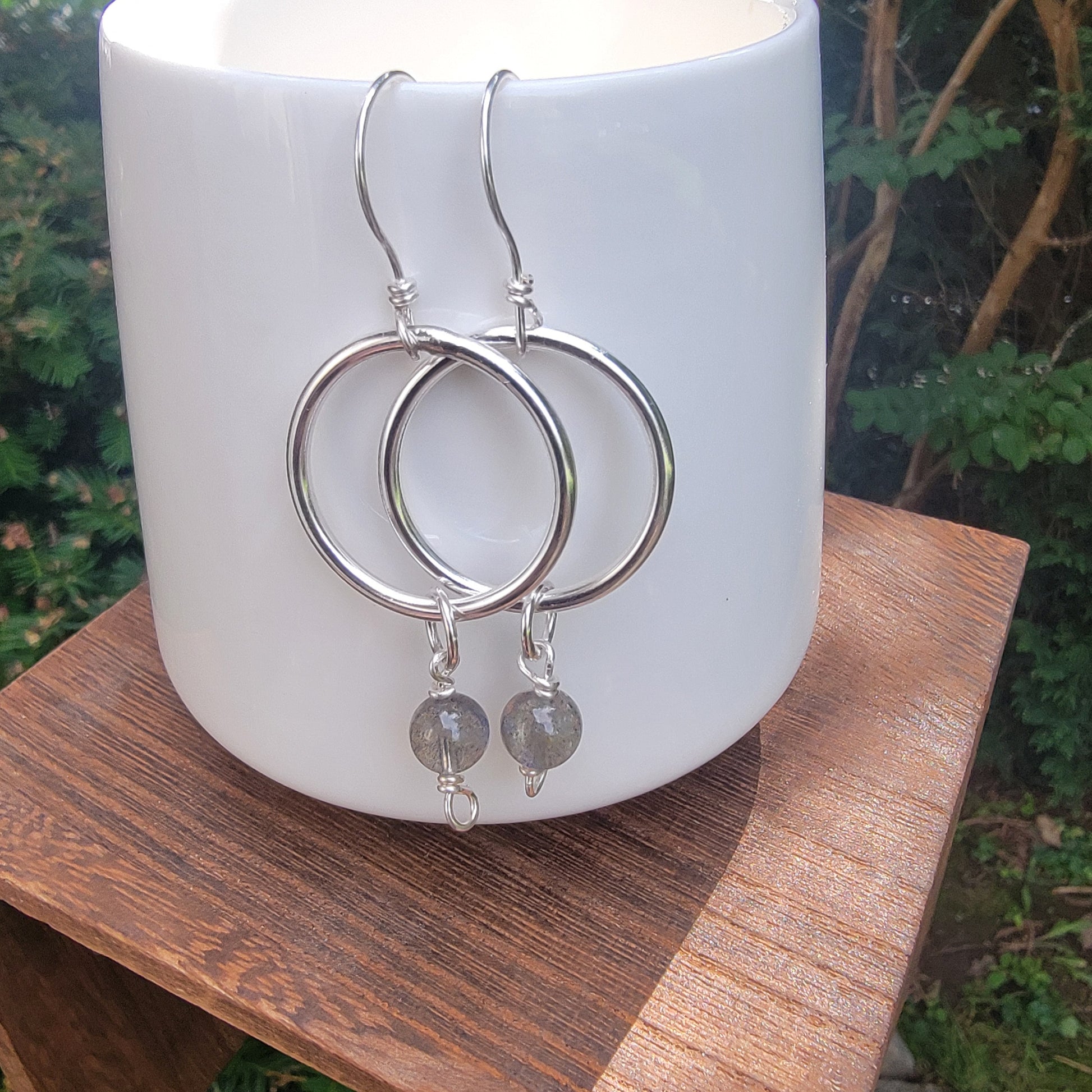 Silver hoop earrings with small gray stone dangles, displayed on a white ceramic candle holder set on a wooden surface with an outdoor garden background.