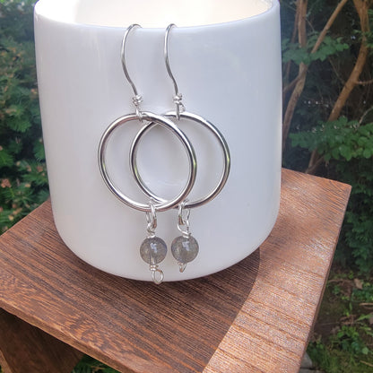Silver hoop earrings with small gray stone dangles, displayed on a white ceramic candle holder set on a wooden surface with an outdoor garden background.