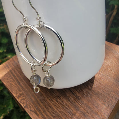 Silver hoop earrings with small gray stone bead drops, displayed on a white ceramic vase resting on a wooden surface.