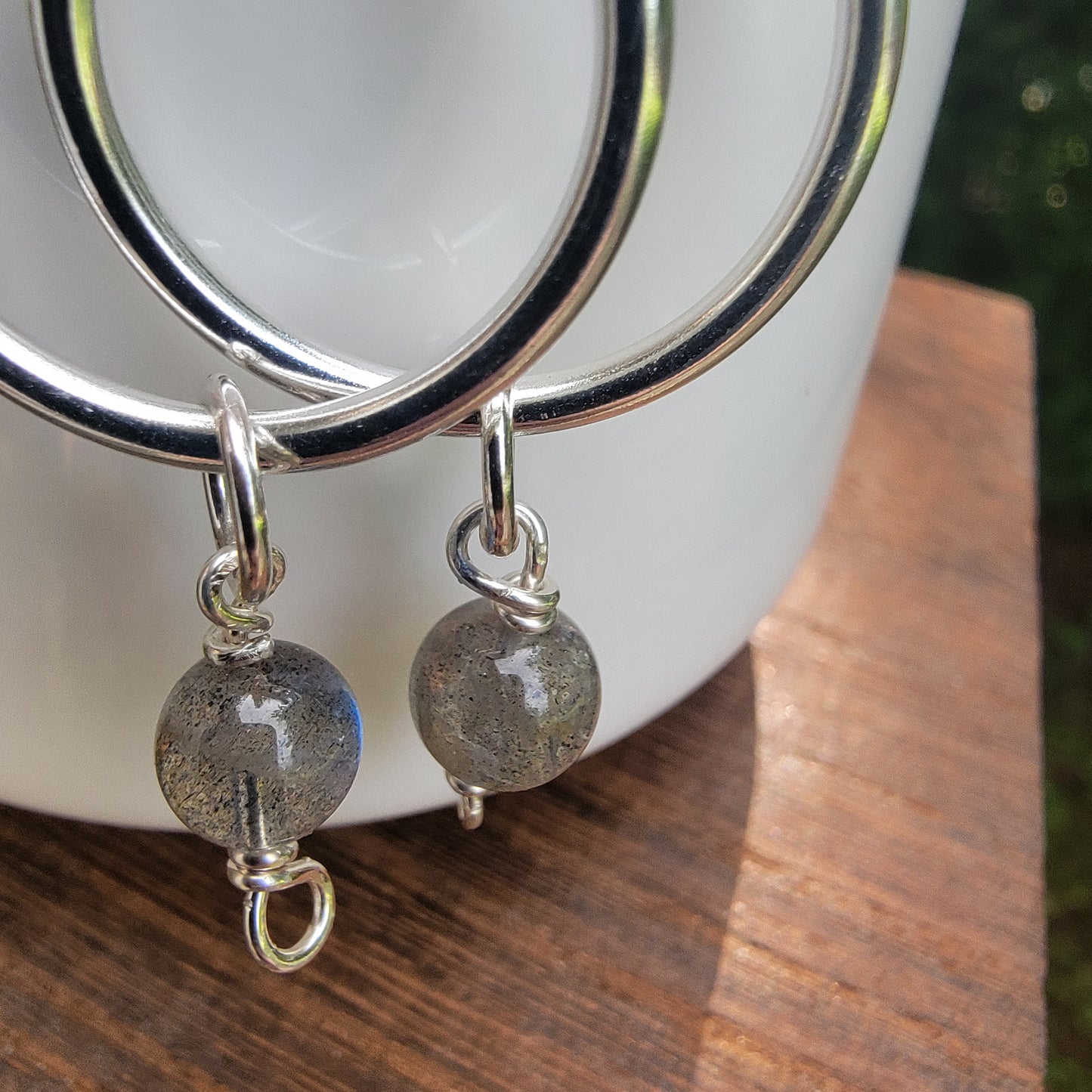 A close-up of a pair of silver hoop earrings with dangling round gray labradorite beads, resting on a white ceramic cup over a wooden surface.