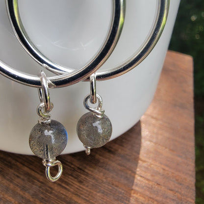 A close-up of a pair of silver hoop earrings with dangling round gray labradorite beads, resting on a white ceramic cup over a wooden surface.
