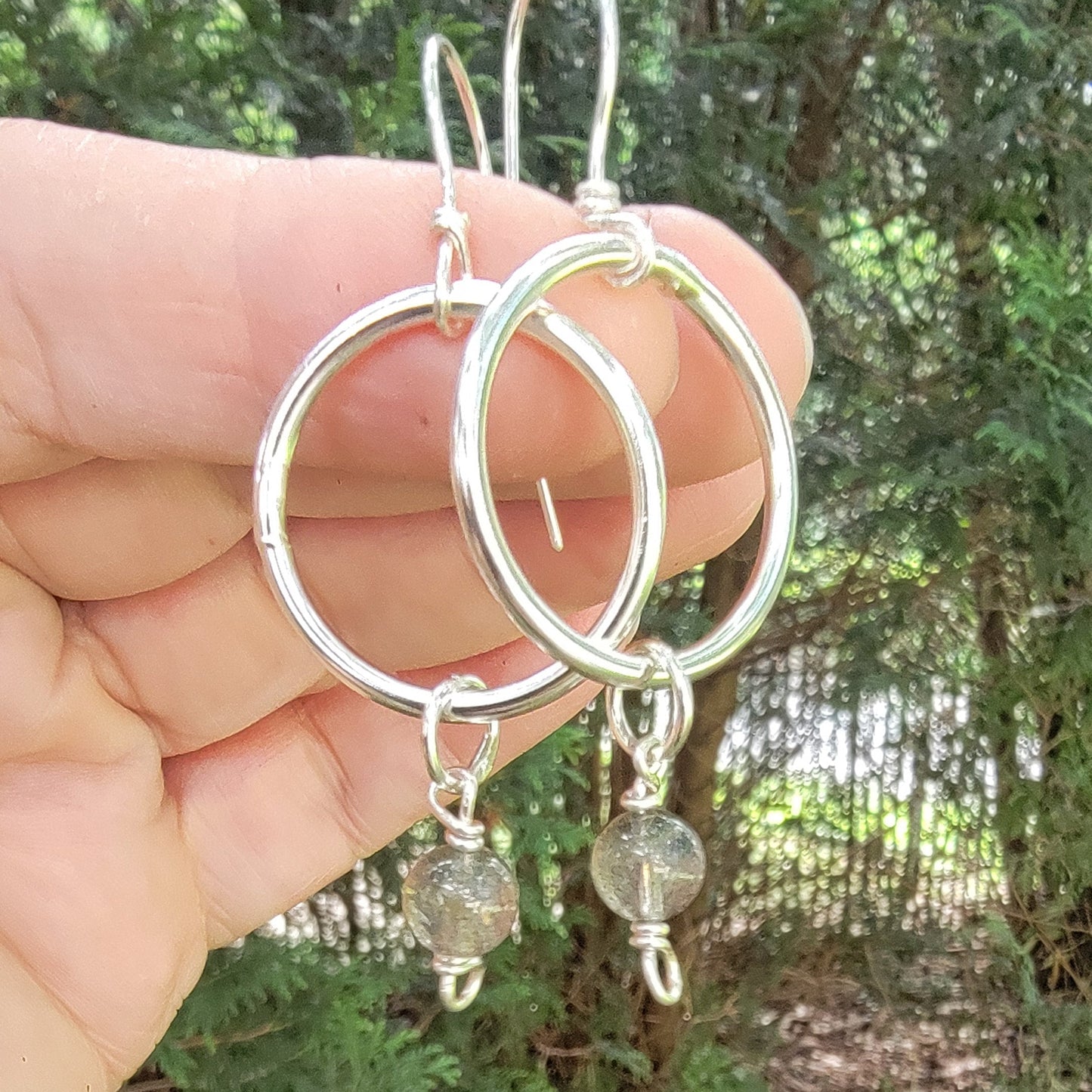 A pair of handmade silver hoop earrings with small clear faceted beads and fishhook wires, held in a person's hand against a blurry background of green foliage and trees.