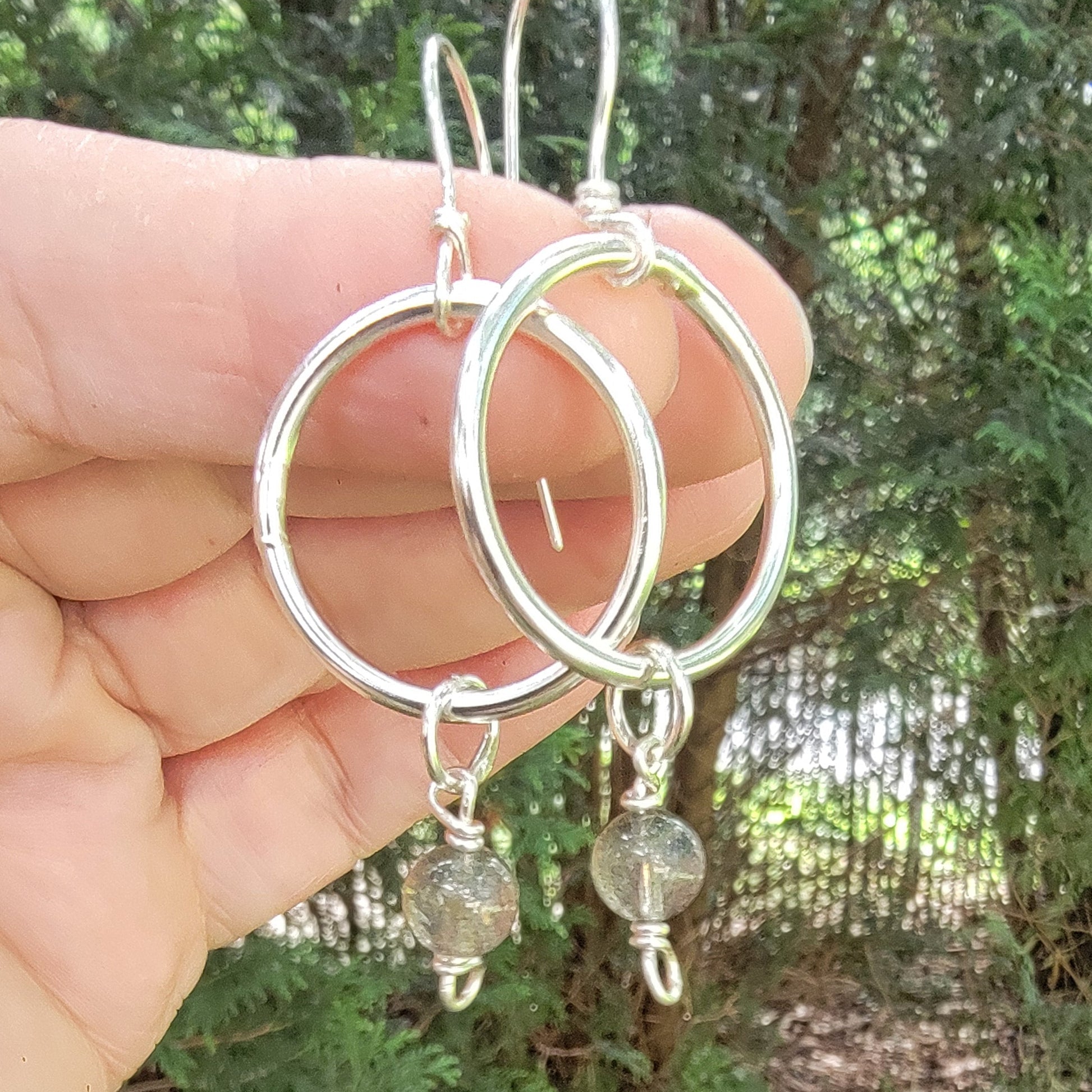 A pair of handmade silver hoop earrings with small clear faceted beads and fishhook wires, held in a person's hand against a blurry background of green foliage and trees.