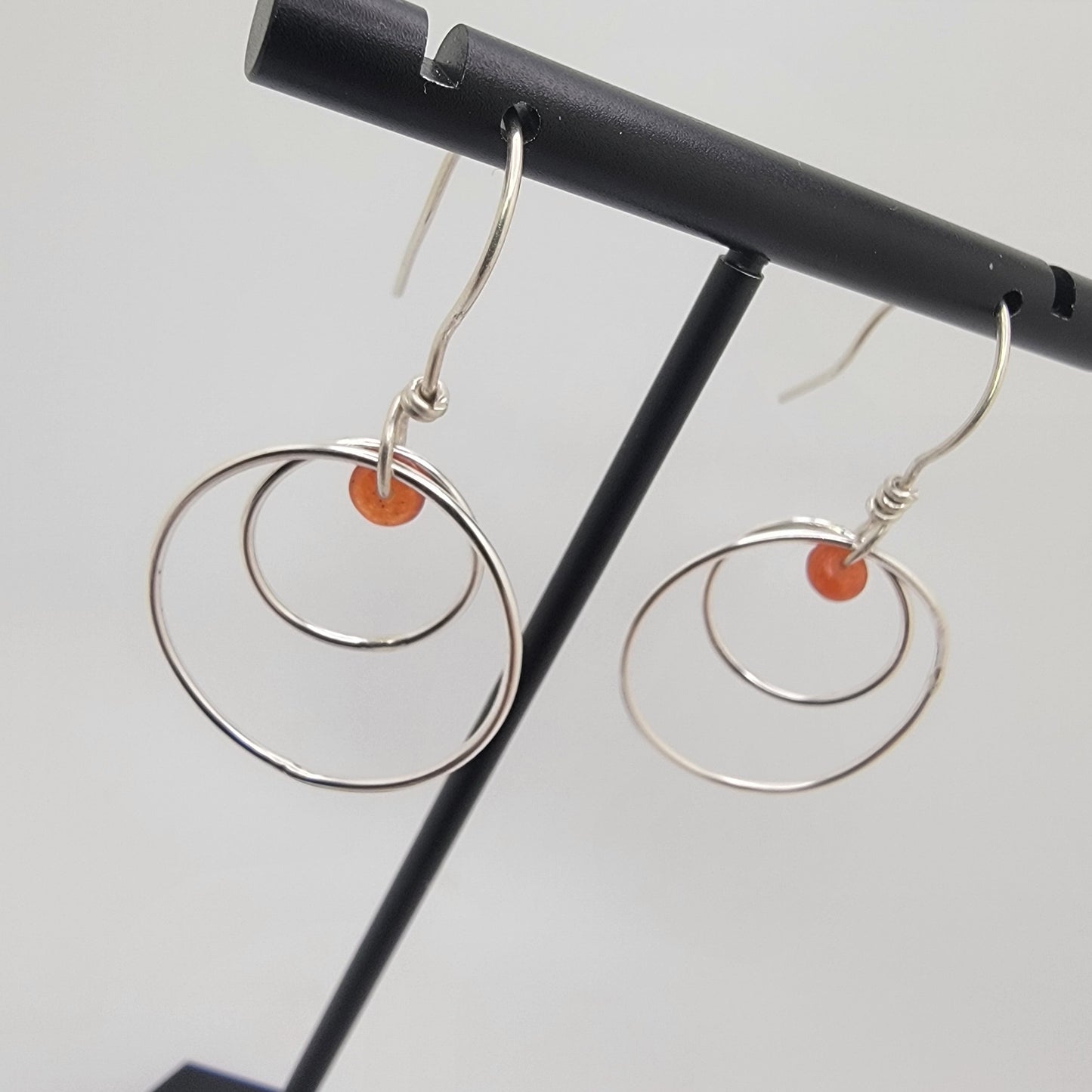 A pair of silver-toned double-ring dangle earrings with small orange bead accents, displayed on a black jewelry stand against a light gray background.