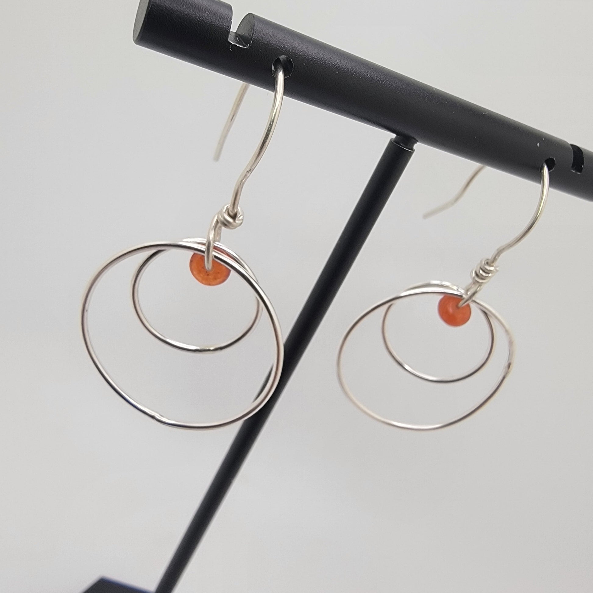 A pair of silver-toned double-ring dangle earrings with small orange bead accents, displayed on a black jewelry stand against a light gray background.
