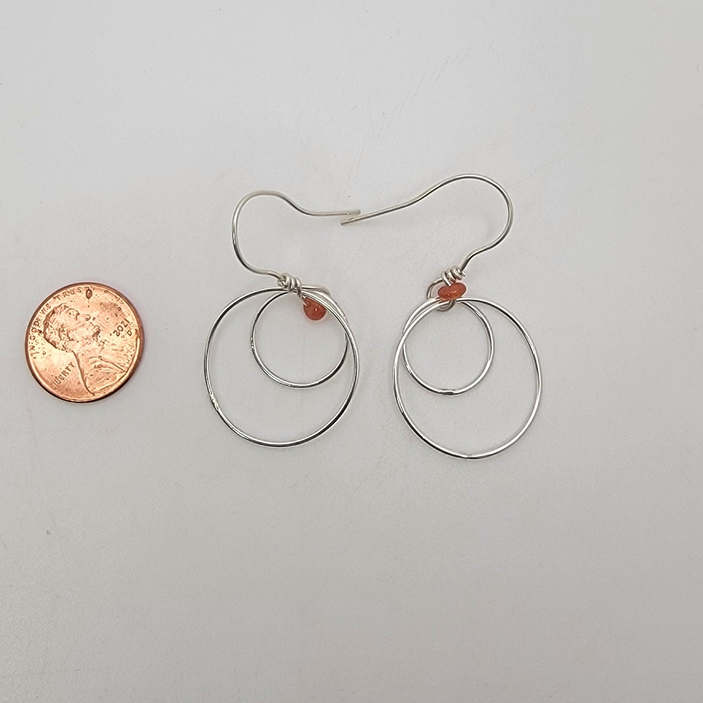 A pair of silver-toned wire hoop earrings with small orange-red bead accents, displayed on a plain light background next to a penny for scale.