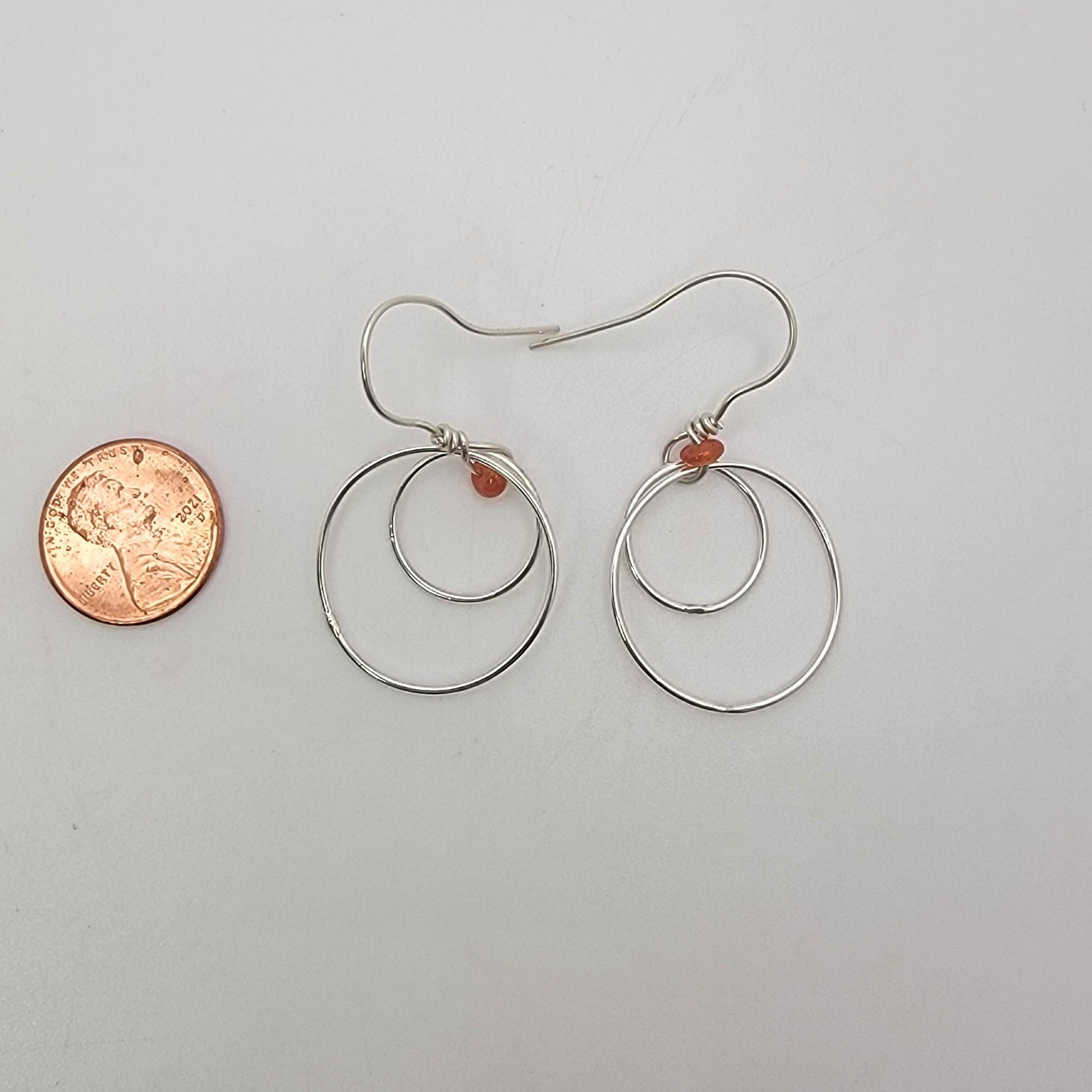 A pair of silver-toned wire hoop earrings with small orange-red bead accents, displayed on a plain light background next to a penny for scale.