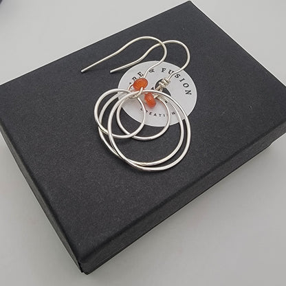 A pair of silver-toned drop earrings with small orange-red bead accents, presented on a white circular card atop a black square box against a light gray background.