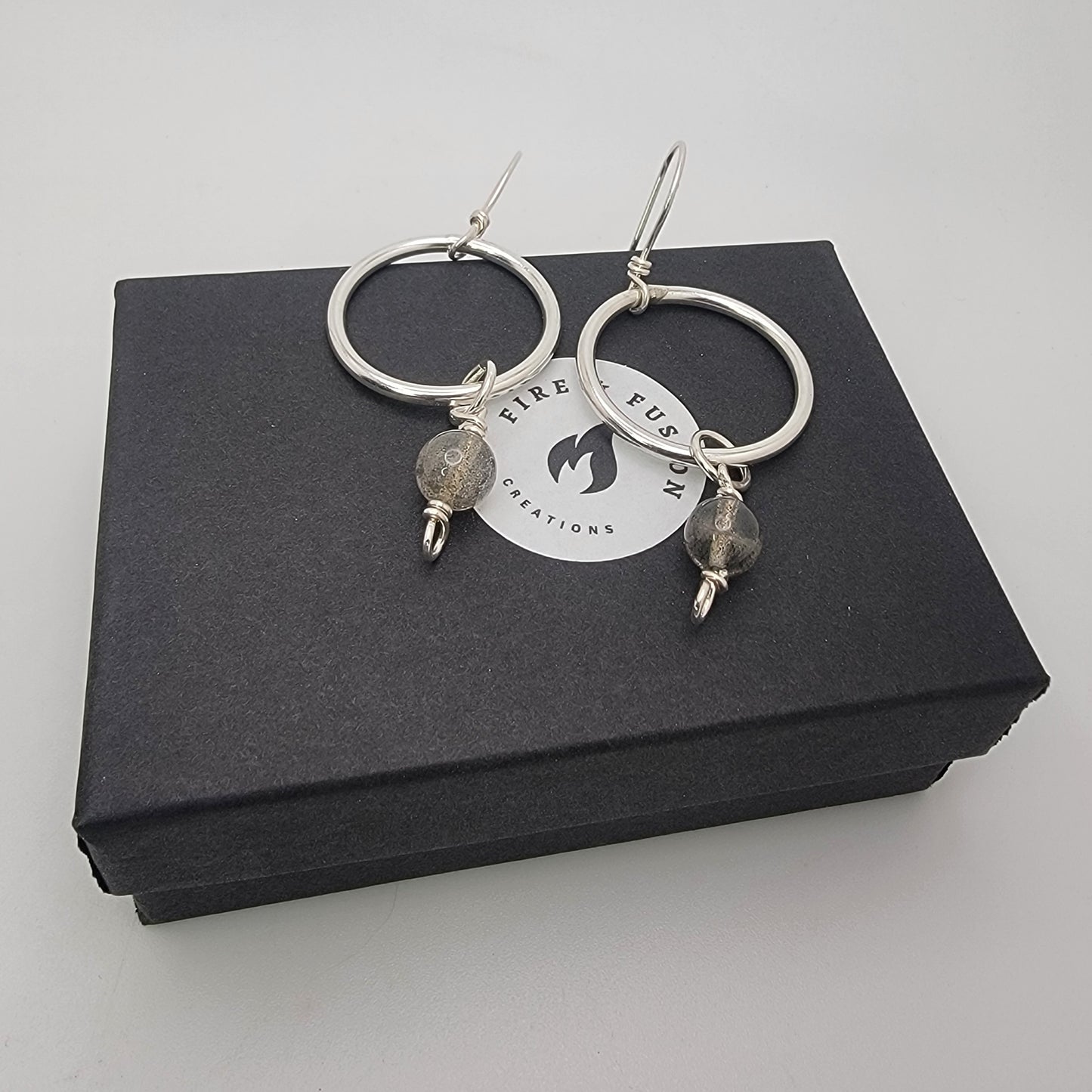 A pair of silver hoop earrings with small gray stone beads hanging from the hoops, displayed on top of a black gift box with a circular Firefly Creations logo.